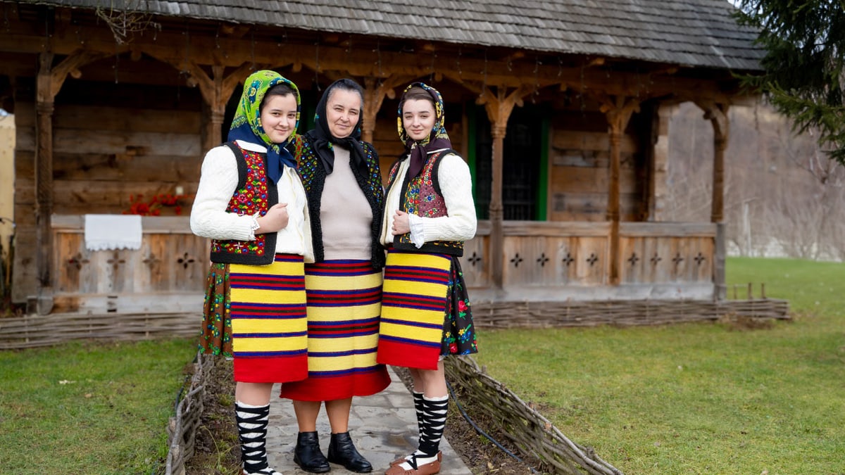 Three women in colorful traditional attire stand outside a wooden house in Maramures, Romania.